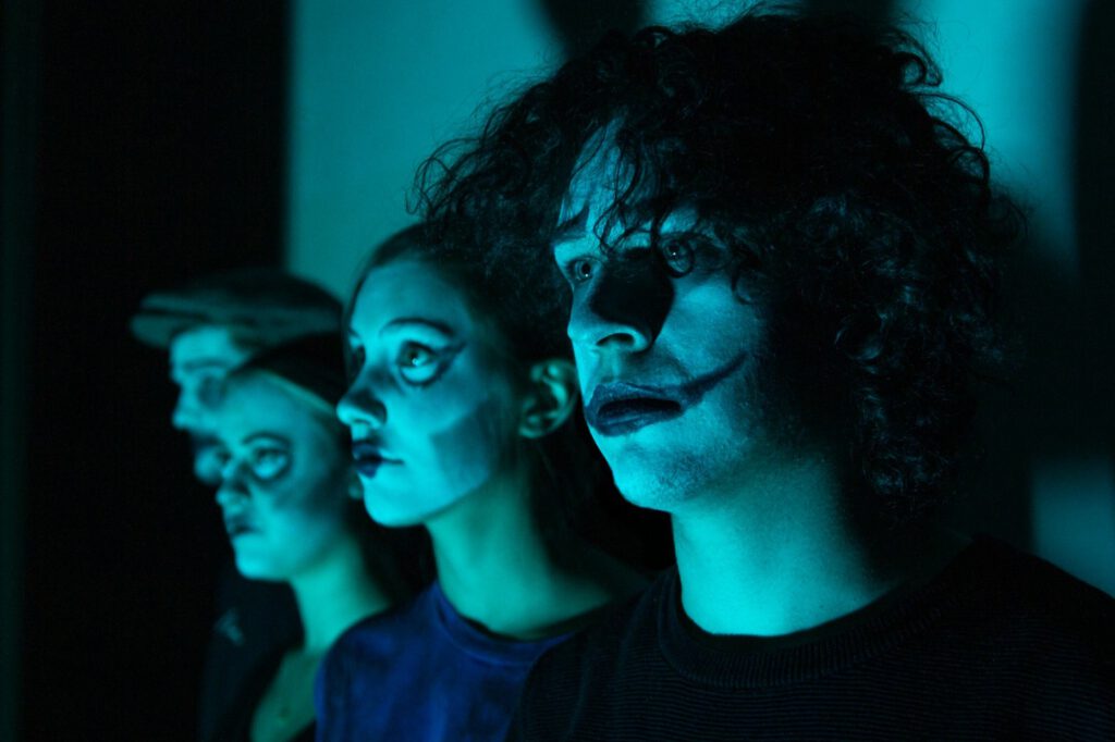 Vier junge Darstellende mit expressiver Schminke und dramatischer Beleuchtung in einer experimentellen Theaterszene.