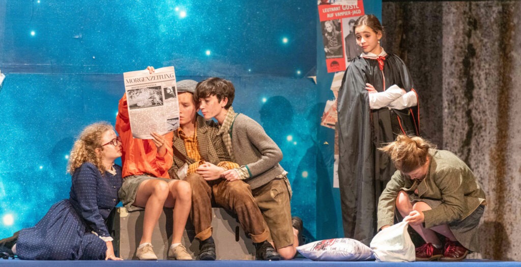 Jugendliche in einer kreativen Theaterszene – Zeitung als Requisite, Sternenhimmel im Hintergrund, Vampirfigur und Vintage-Plakate als Teil der Kulisse.